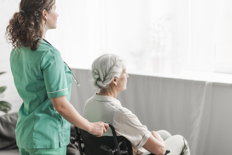 Infirmière à domicile en blouse verte soutenant une patiente âgée en fauteuil roulant regardant vers la fenêtre